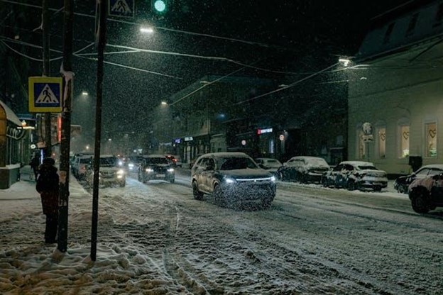 snowy urban street at night showing cars driving on a snow-covered road